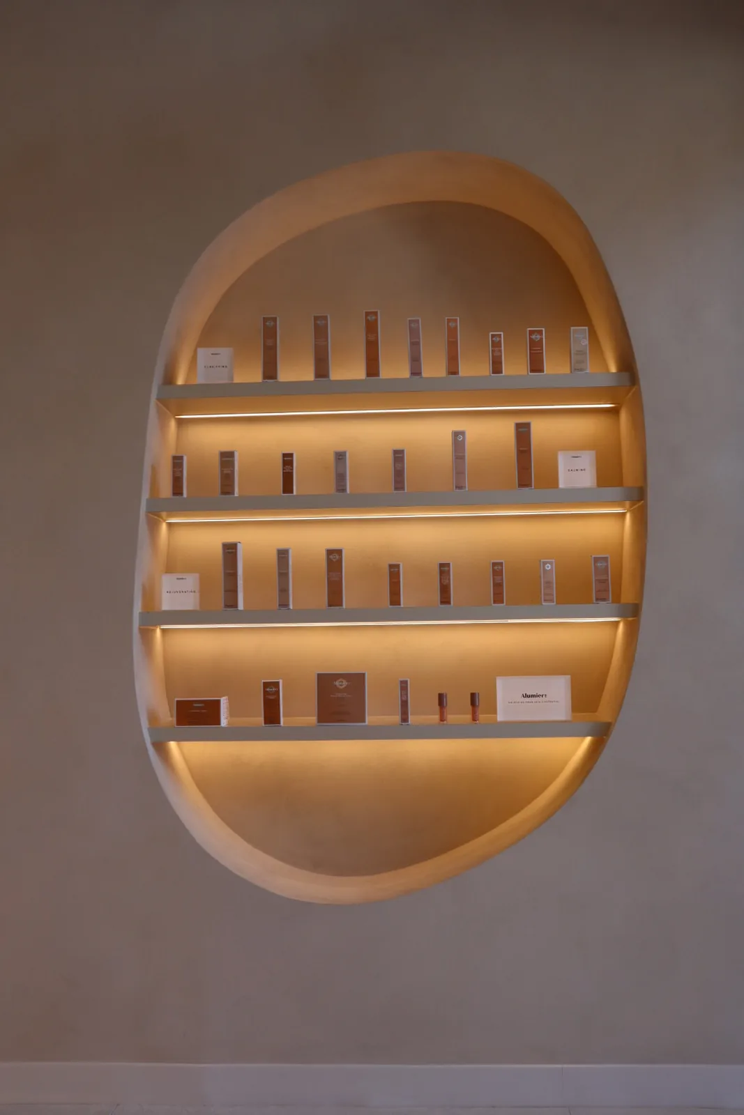 Oval retail display niche carved into a two-tone limewash feature wall with backlit shelves of skincare products, Envy Cosmetic Clinic Ottawa
